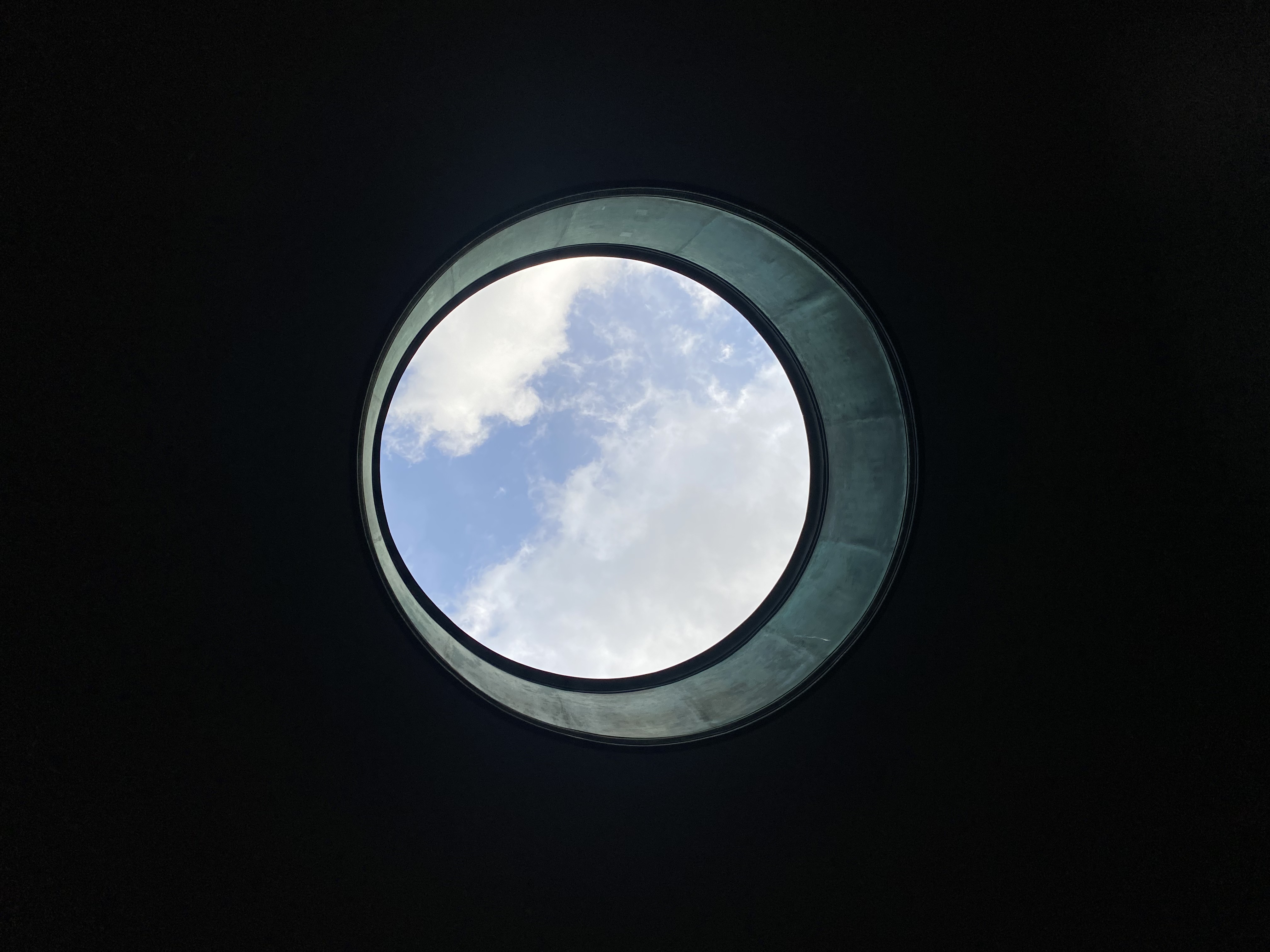 A circular oculus aperture cut into dark concrete, framing a disc of blue sky with white cloud